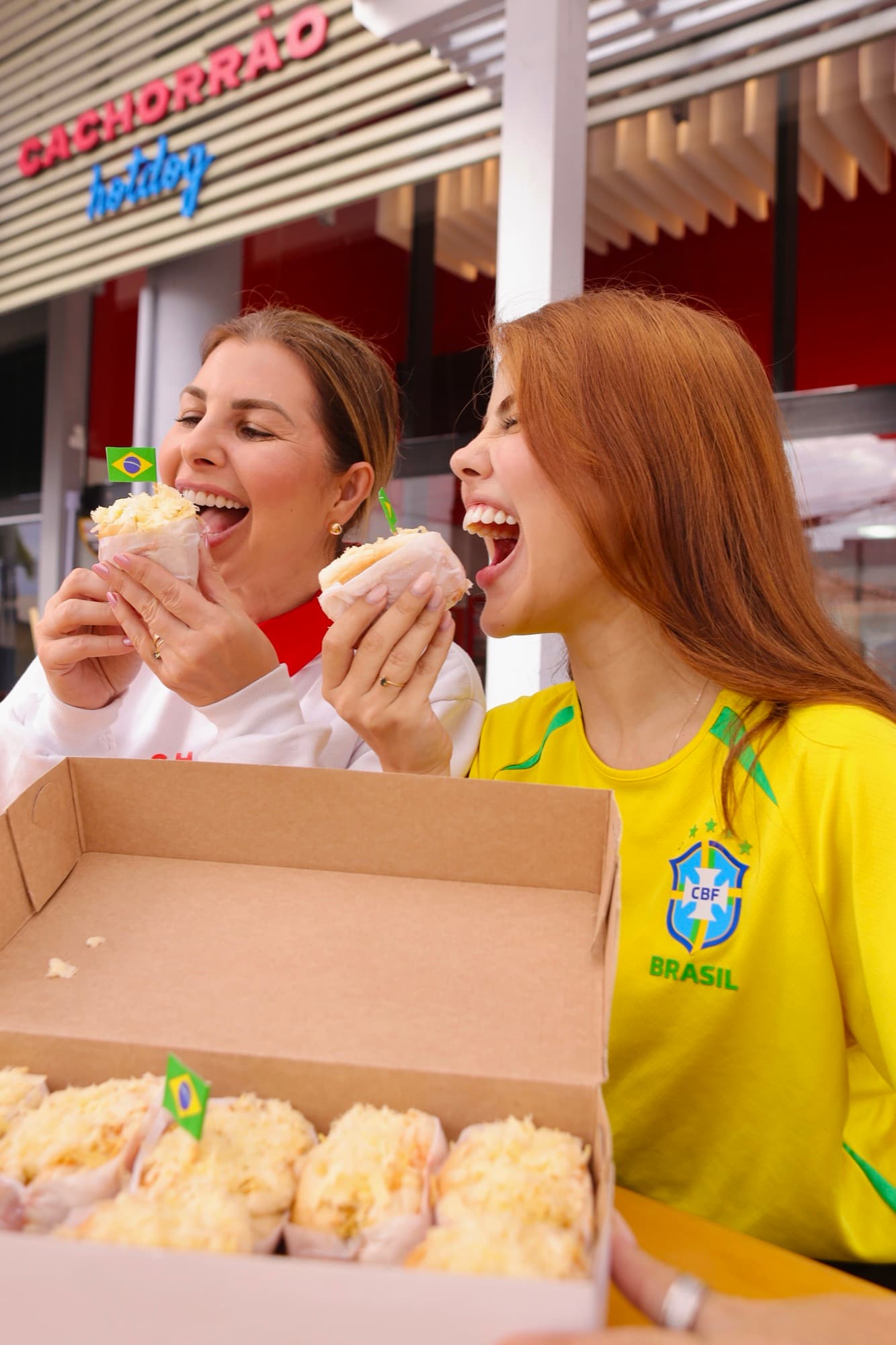 Amigas comendo mini-dogs ao lado de uma caixa cheia, prontos para encomenda